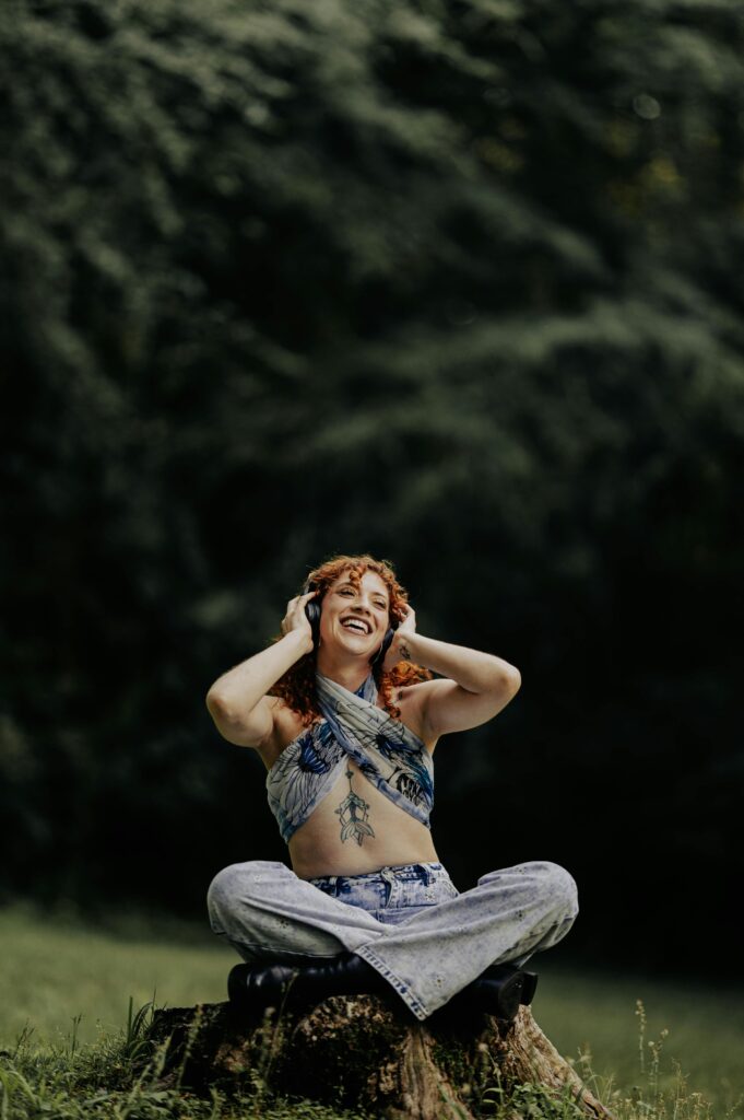 Redheaded woman sitting on a tree stump, enjoying music in a lush forest setting.