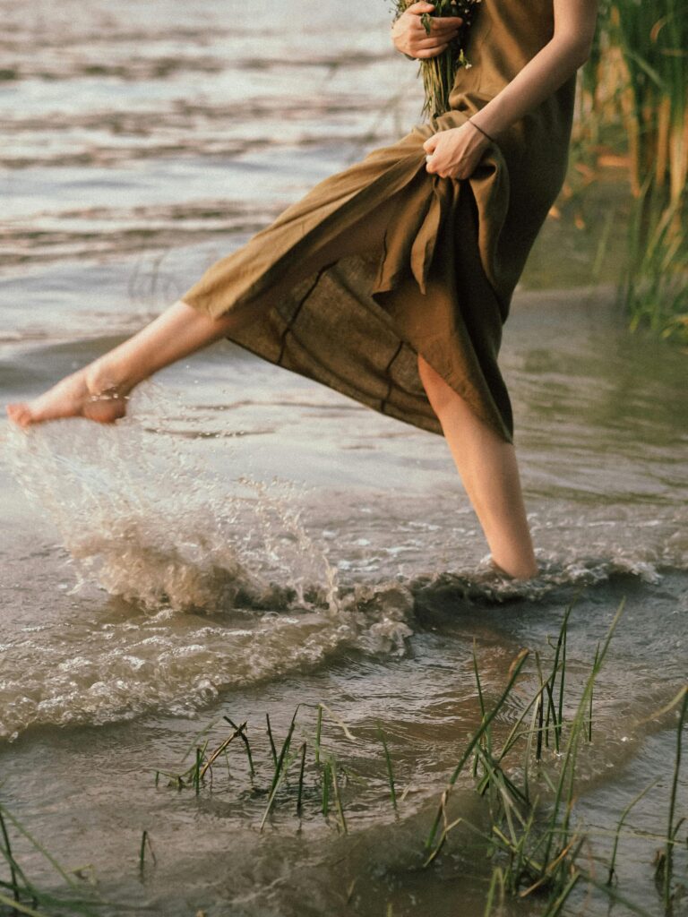 A woman in a dress playfully splashing water while standing barefoot by a lake.